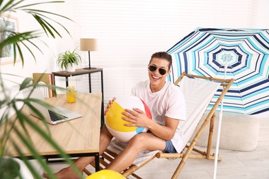 Photo of Happy man in sunglasses with beach ball having fun on sunbed at table indoors. Dreaming about vacation at sea
