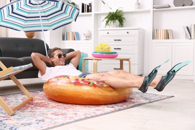 Photo of Happy man in sunglasses and flippers with inflatable ring having fun on floor at home. Dreaming about vacation at sea