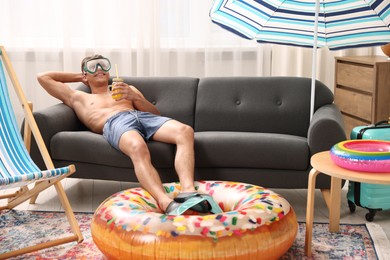 Photo of Happy man in diving mask and flippers with mason jar of juice and inflatable ring on sofa at home. Dreaming about vacation at sea