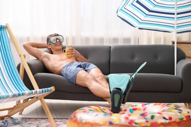 Photo of Happy man in diving mask and flippers with mason jar of juice and inflatable ring on sofa at home. Dreaming about vacation at sea