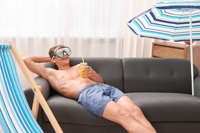 Photo of Happy man in diving mask with mason jar of juice on sofa at home. Dreaming about vacation at sea