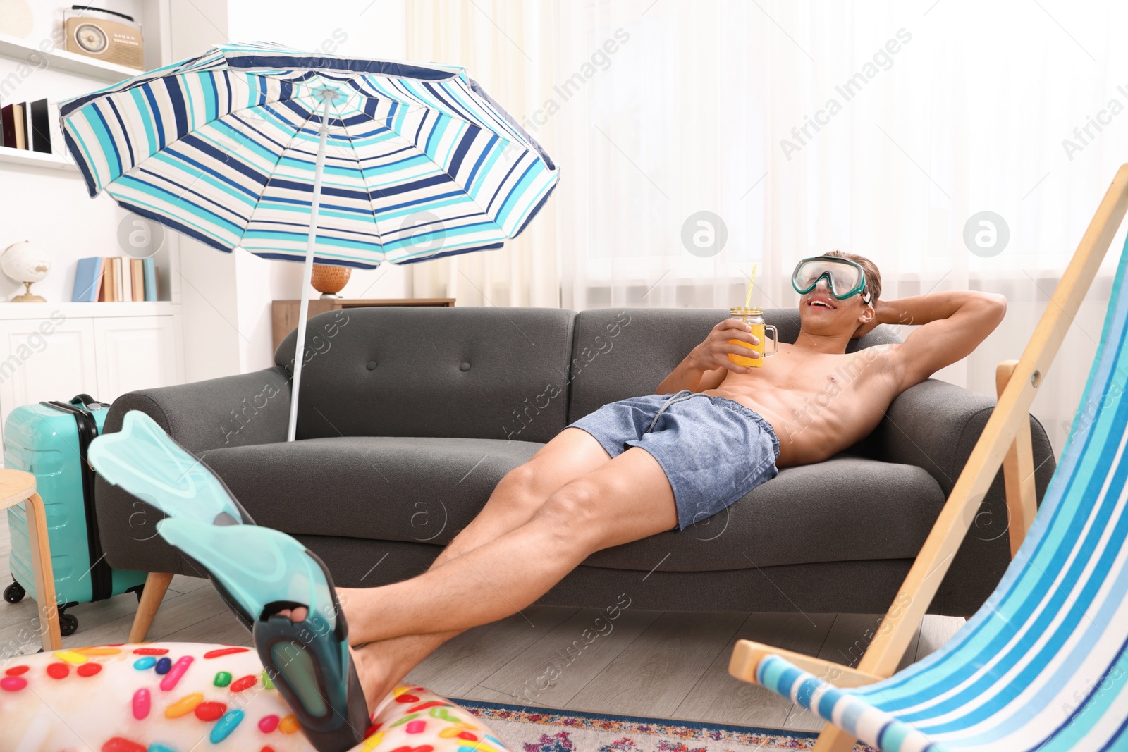 Happy man in diving mask and flippers with mason jar of juice on sofa at home. Dreaming about vacation at sea Photo of Happy man in diving mask and flippers with mason jar of juice on sofa at home. Dreaming about vacation at sea