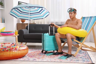 Photo of Man in flippers with diving mask, suitcase and inflatable ring on folding chair at home. Dreaming about vacation at sea