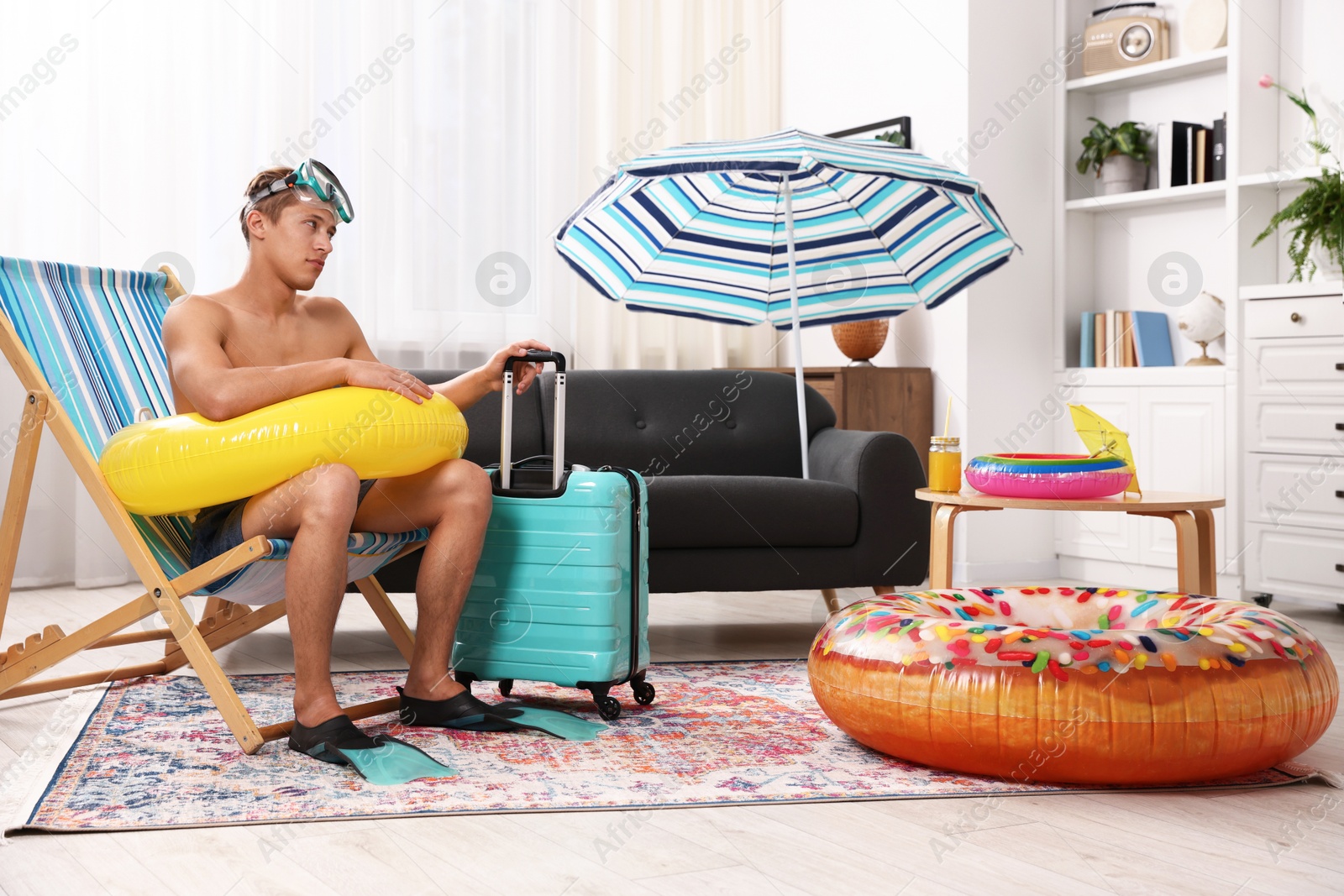 Man in flippers with diving mask, suitcase and inflatable ring on folding chair at home. Dreaming about vacation at sea Photo of Man in flippers with diving mask, suitcase and inflatable ring on folding chair at home. Dreaming about vacation at sea