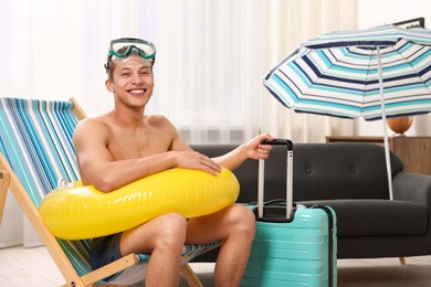 Photo of Happy man with diving mask, suitcase and inflatable ring on folding chair at home. Dreaming about vacation at sea