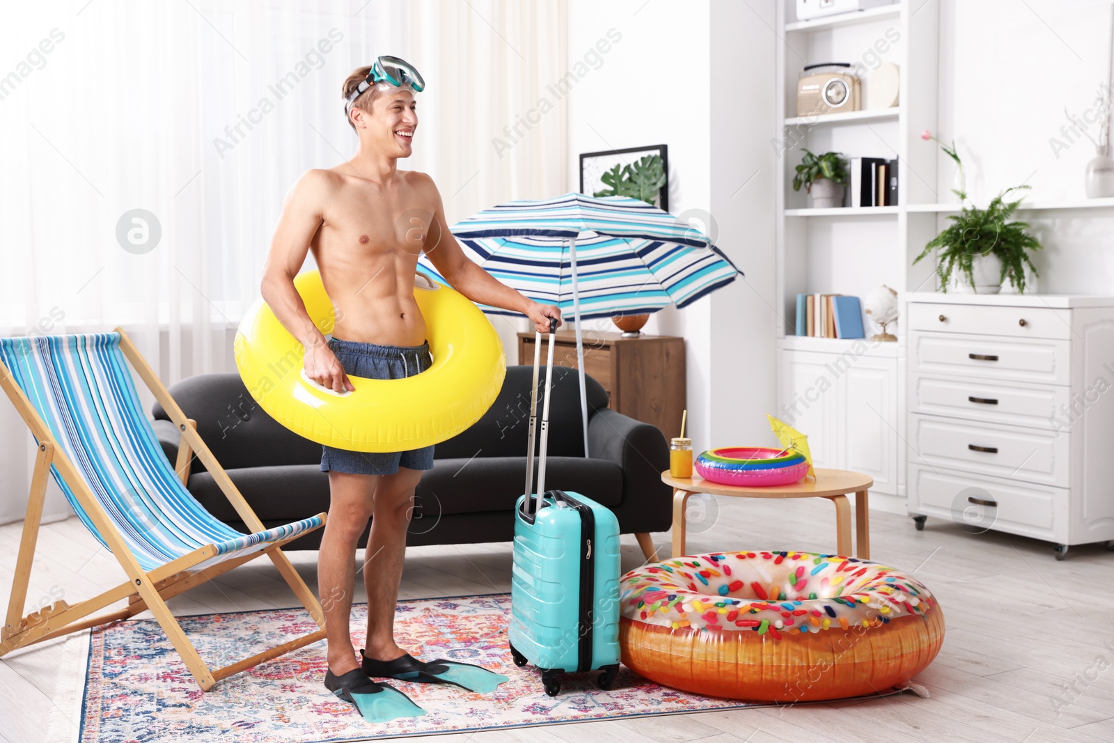 Happy man in flippers with diving mask, suitcase and inflatable ring at home. Dreaming about vacation at sea Photo of Happy man in flippers with diving mask, suitcase and inflatable ring at home. Dreaming about vacation at sea