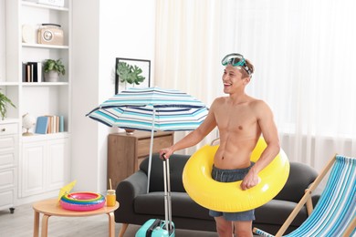 Photo of Happy man with diving mask, suitcase and inflatable ring at home. Dreaming about vacation at sea