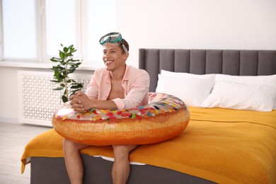 Photo of Happy man with diving mask and inflatable ring on bed at home. Dreaming about vacation at sea