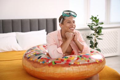 Photo of Happy man with diving mask and inflatable ring on bed at home. Dreaming about vacation at sea