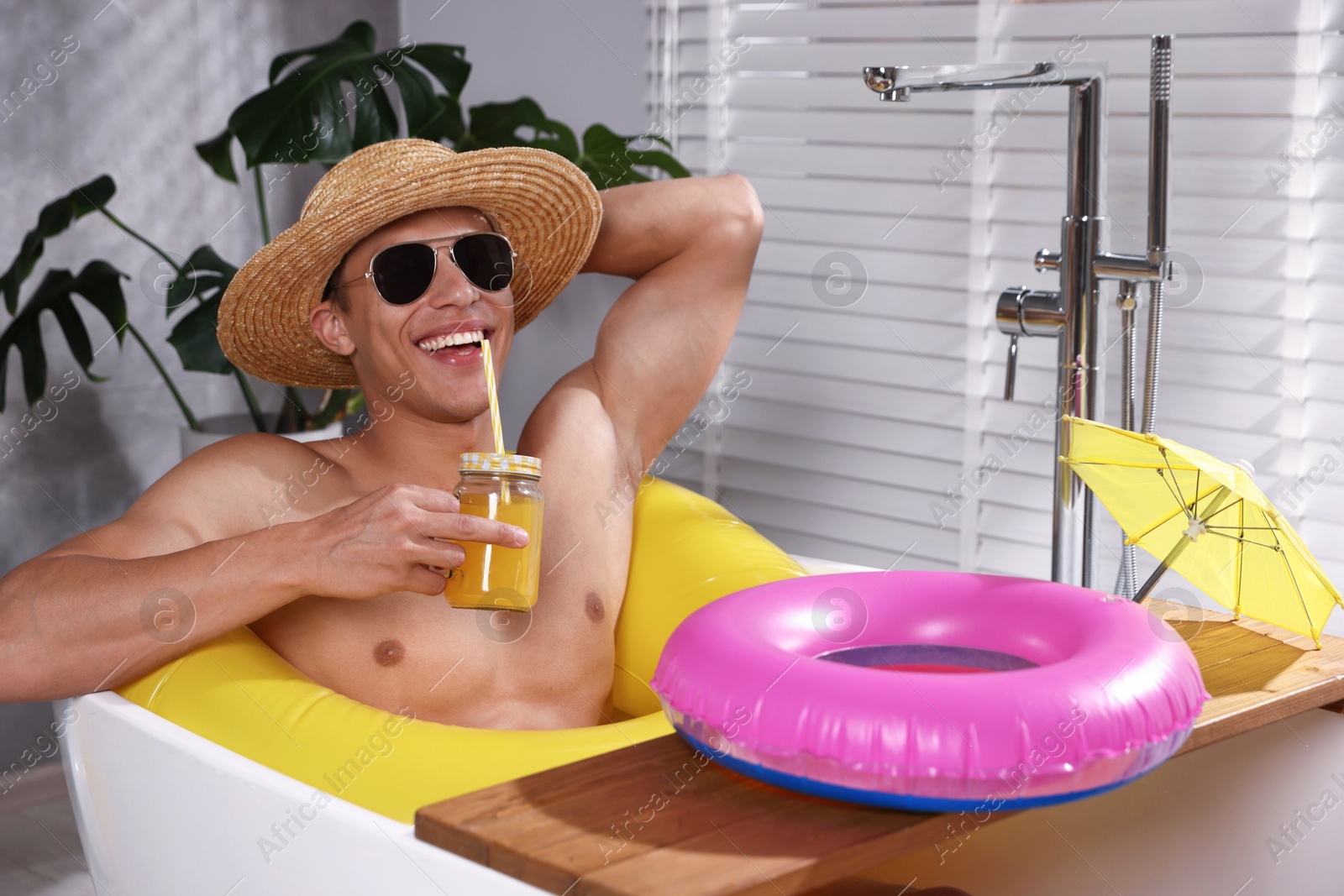 Photo of Happy man in sunglasses and straw hat drinking juice from mason jar with inflatable ring in bathtub at home. Dreaming about vacation at sea