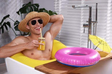 Photo of Happy man in sunglasses and straw hat drinking juice from mason jar with inflatable ring in bathtub at home. Dreaming about vacation at sea