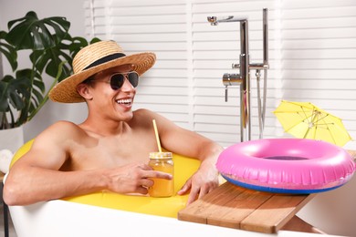 Photo of Happy man in sunglasses and straw hat drinking juice from mason jar with inflatable ring in bathtub at home. Dreaming about vacation at sea