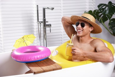 Photo of Happy man in sunglasses and straw hat drinking juice from mason jar with inflatable ring in bathtub at home. Dreaming about vacation at sea