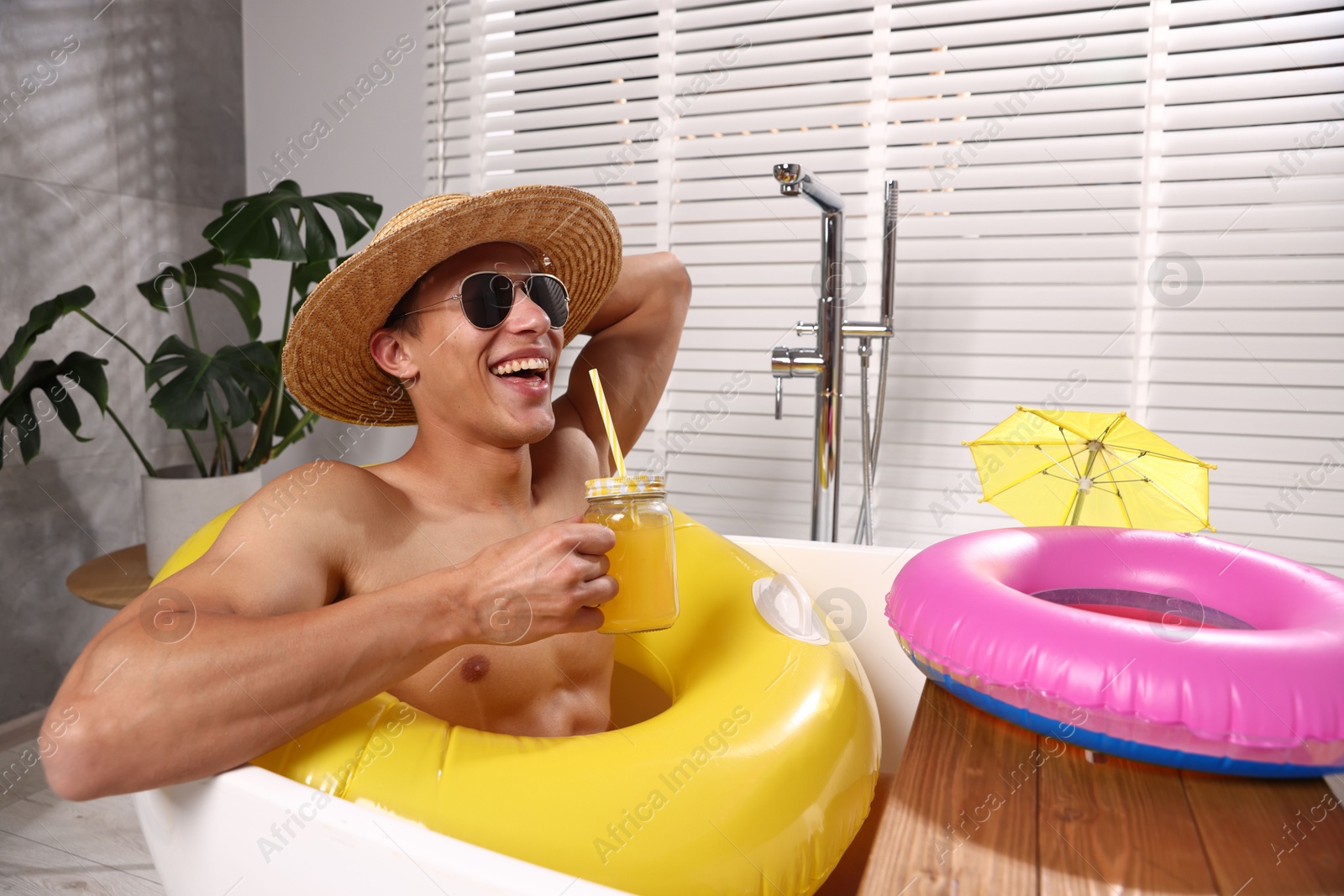 Happy man in sunglasses and straw hat drinking juice from mason jar with inflatable ring in bathtub at home. Dreaming about vacation at sea Photo of Happy man in sunglasses and straw hat drinking juice from mason jar with inflatable ring in bathtub at home. Dreaming about vacation at sea
