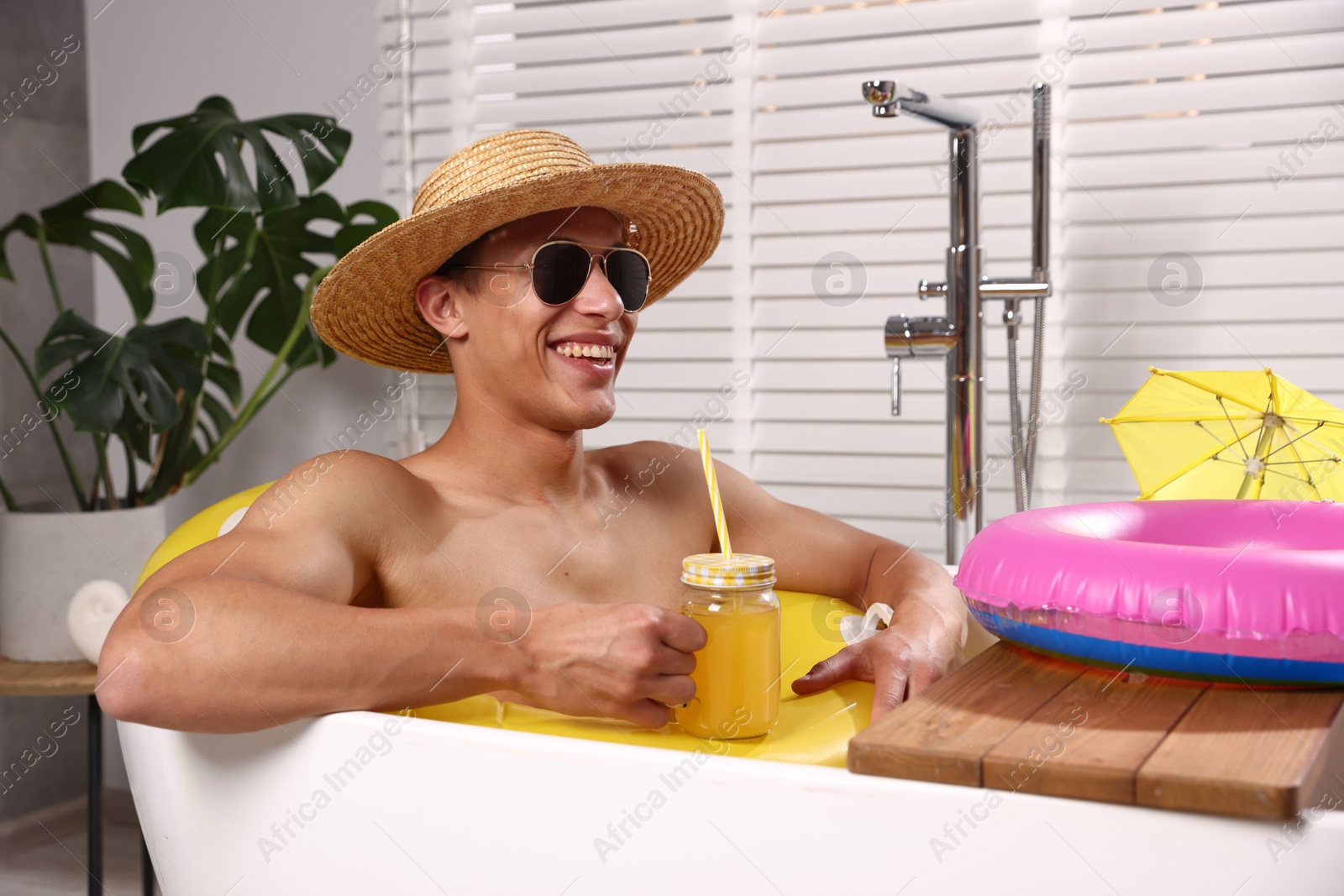 Happy man in sunglasses and straw hat drinking juice from mason jar with inflatable ring in bathtub at home. Dreaming about vacation at sea Photo of Happy man in sunglasses and straw hat drinking juice from mason jar with inflatable ring in bathtub at home. Dreaming about vacation at sea