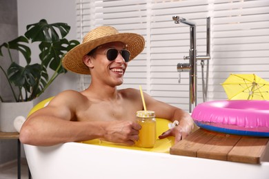 Photo of Happy man in sunglasses and straw hat drinking juice from mason jar with inflatable ring in bathtub at home. Dreaming about vacation at sea