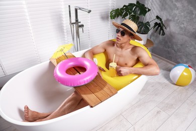 Photo of Young man in sunglasses and straw hat drinking juice from mason jar with inflatable ring in bathtub at home. Dreaming about vacation at sea
