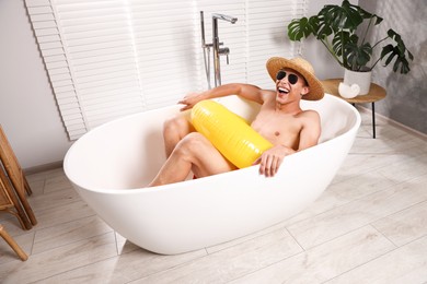 Photo of Happy man in sunglasses and straw hat with inflatable ring chilling in bathtub at home. Dreaming about vacation at sea