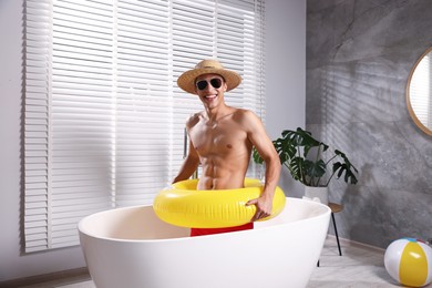 Photo of Happy man in sunglasses and straw hat with inflatable ring chilling in bathtub at home. Dreaming about vacation at sea