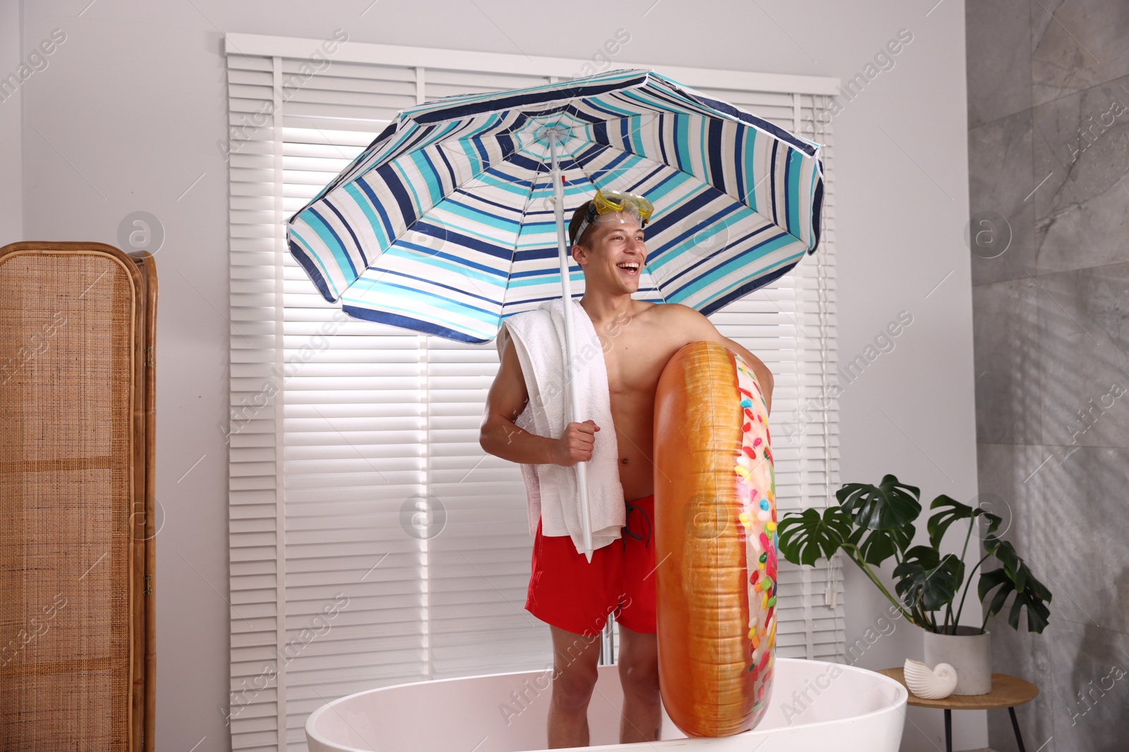 Happy man in with inflatable ring, diving mask, towel and beach umbrella in bathtub at home. Dreaming about vacation at sea Photo of Happy man in with inflatable ring, diving mask, towel and beach umbrella in bathtub at home. Dreaming about vacation at sea