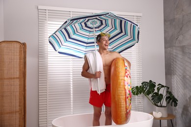 Photo of Happy man in with inflatable ring, diving mask, towel and beach umbrella in bathtub at home. Dreaming about vacation at sea