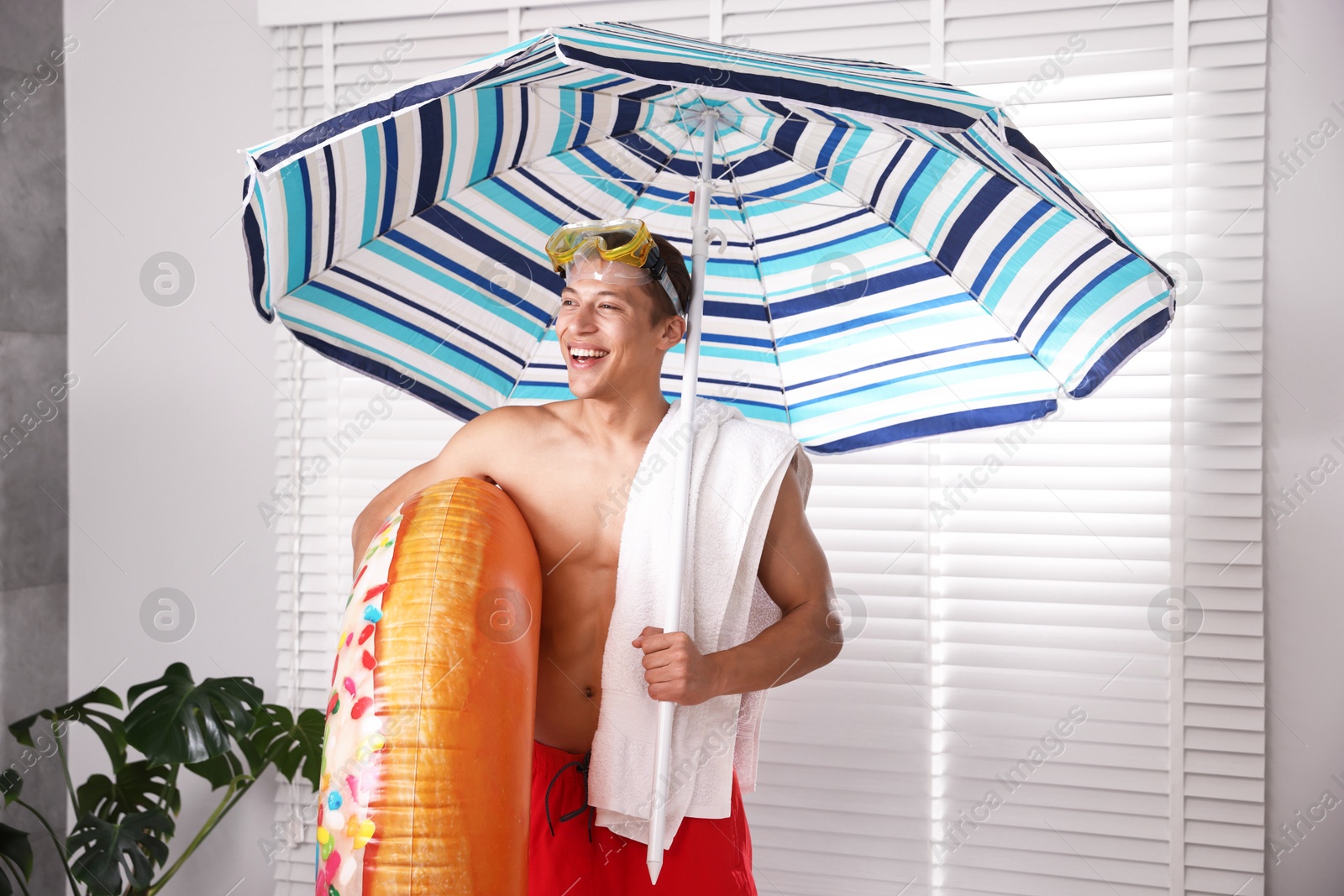 Happy man in with inflatable ring, diving mask, towel and beach umbrella at home. Dreaming about vacation at sea Photo of Happy man in with inflatable ring, diving mask, towel and beach umbrella at home. Dreaming about vacation at sea