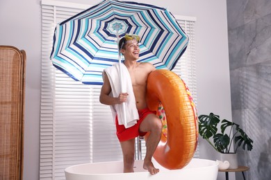 Photo of Happy man in with inflatable ring, diving mask, towel and beach umbrella in bathtub at home. Dreaming about vacation at sea