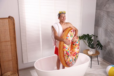 Photo of Happy man in with inflatable ring, diving mask and towel in bathtub at home. Dreaming about vacation at sea