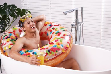 Photo of Happy man with diving mask, mason jar of juice and inflatable ring chilling in bathtub at home. Dreaming about vacation at sea