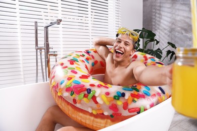 Photo of Happy man with diving mask, mason jar of juice and inflatable ring chilling in bathtub at home. Dreaming about vacation at sea