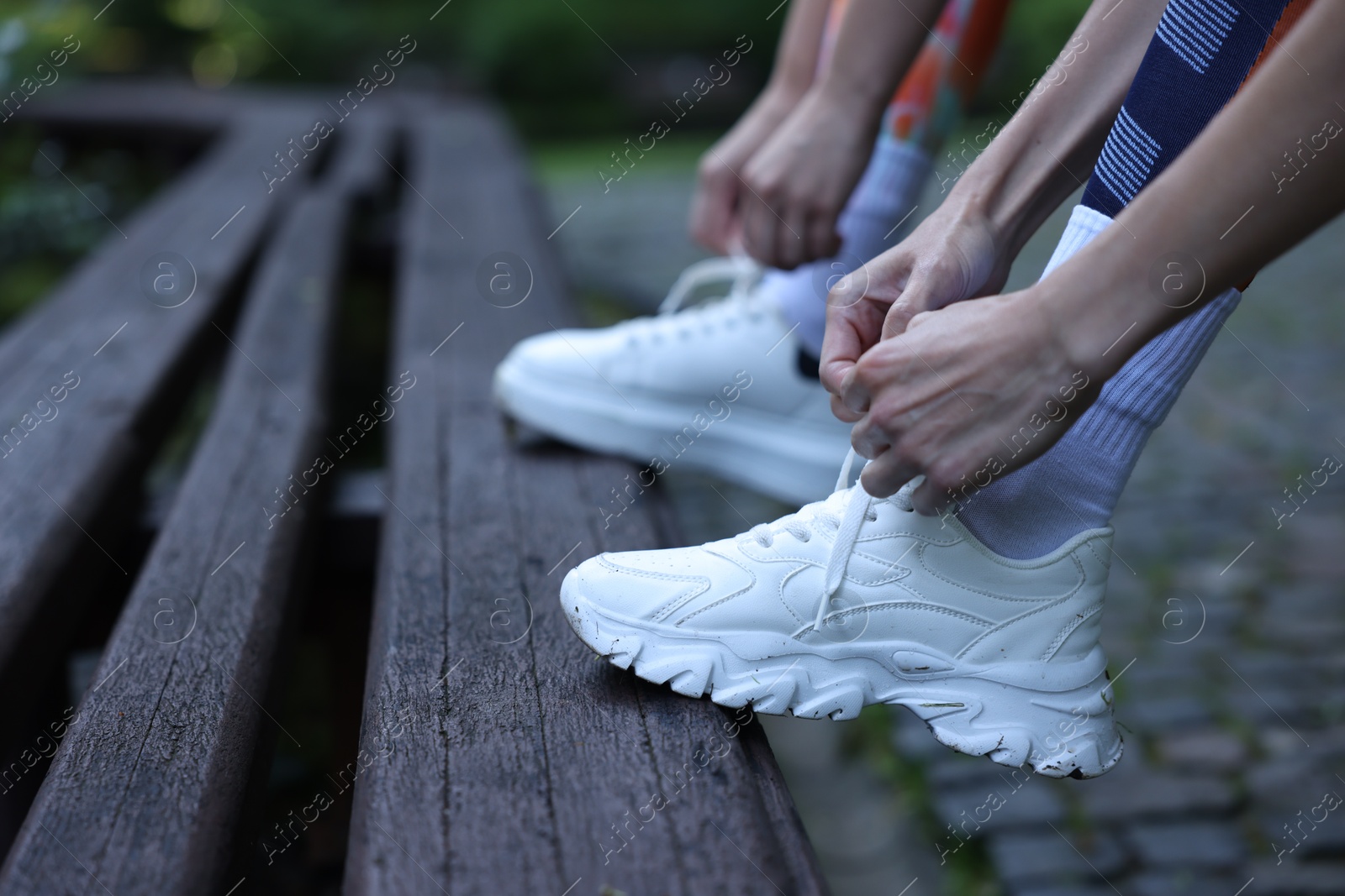 Photo of Young woman and professional personal trainer tying shoes on wooden bench outdoors, closeup. Space for text