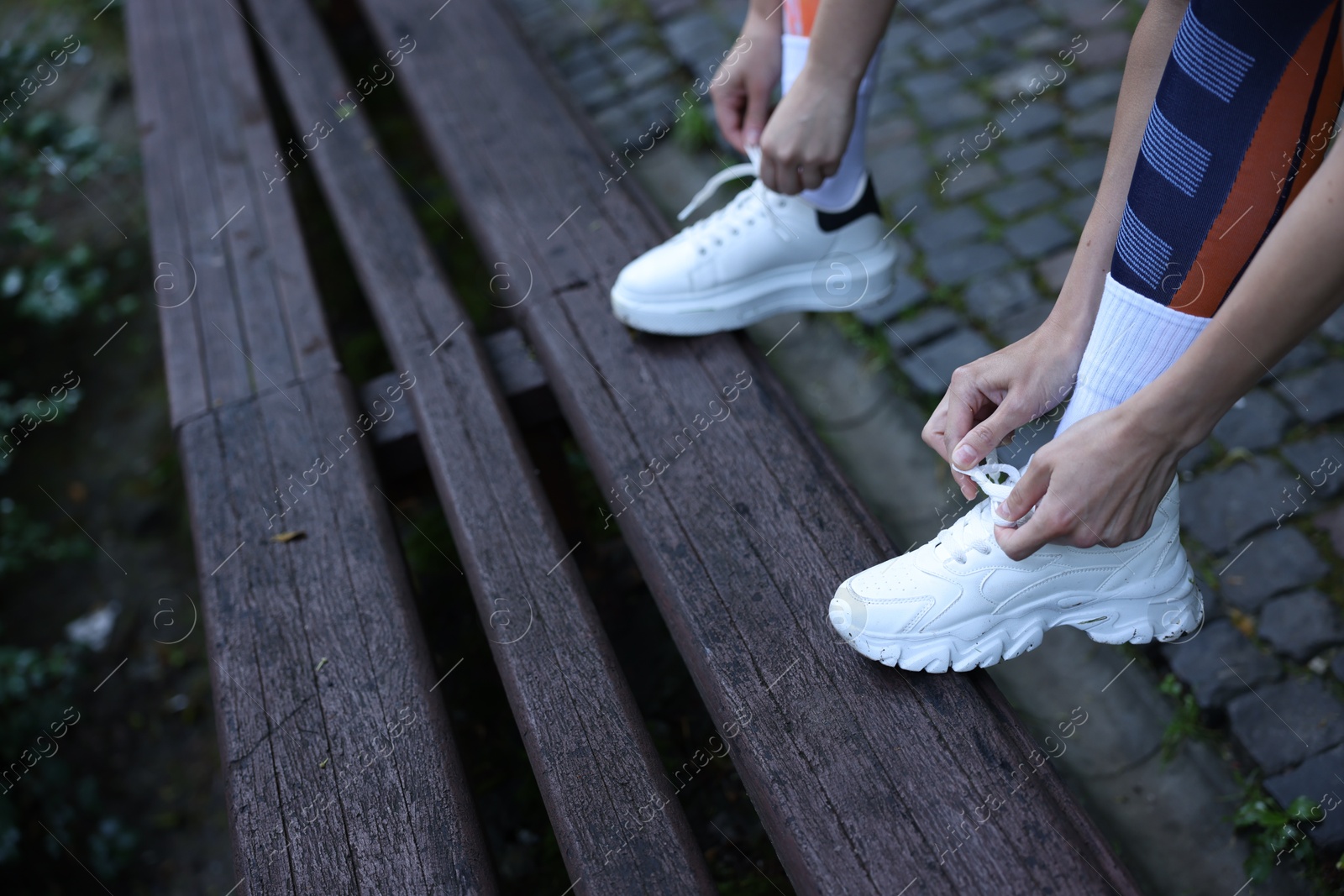 Photo of Young woman and professional personal trainer tying shoes on wooden bench outdoors, closeup. Space for text
