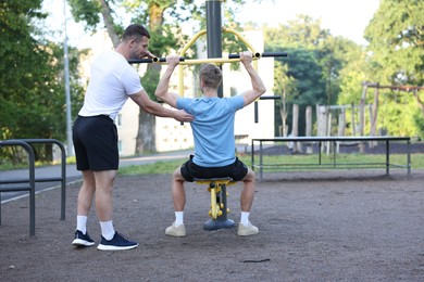 Photo of Young man exercising with outdoor gym equipment with professional personal trainer outdoors, back view. Space for text