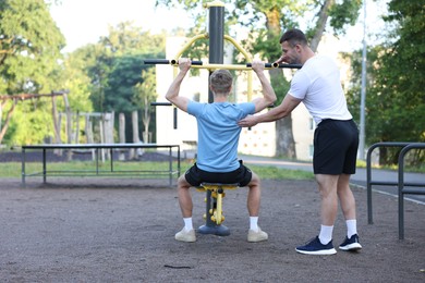 Photo of Young man exercising with outdoor gym equipment with professional personal trainer outdoors, back view. Space for text