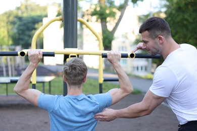 Photo of Young man exercising with outdoor gym equipment with professional personal trainer outdoors, back view