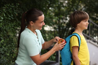 Photo of Mother putting apple into her son's backpack while taking him to school outdoors