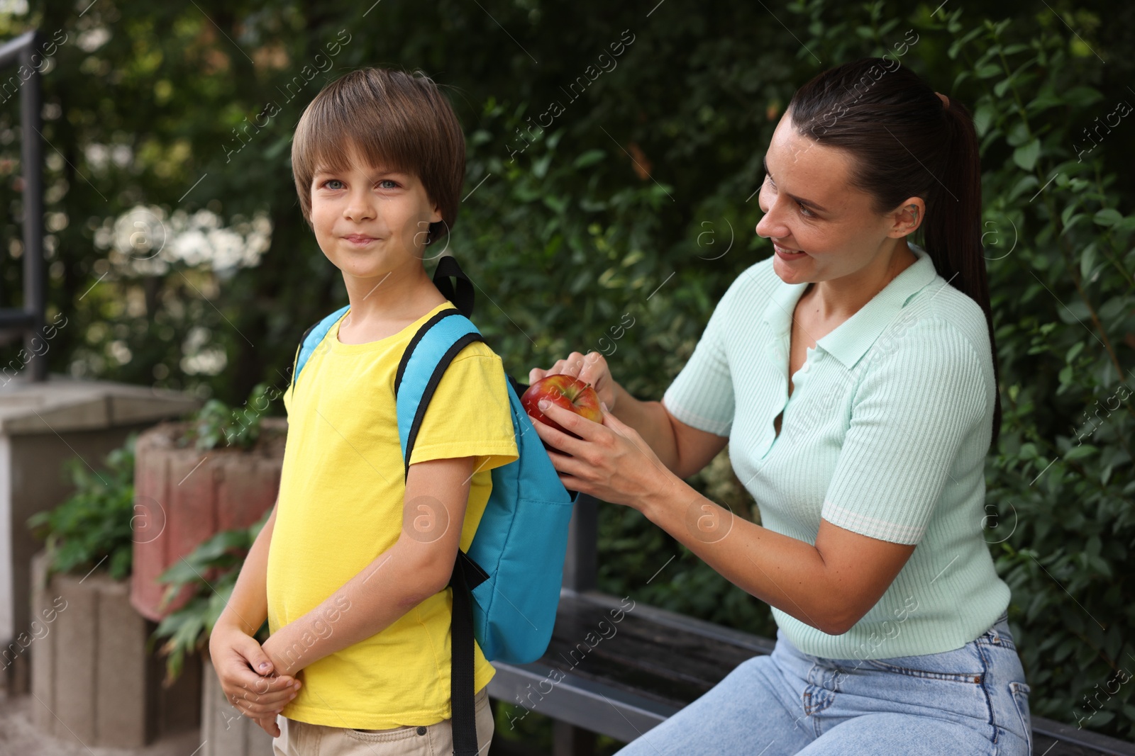 Photo of Mother putting apple into her son's backpack while taking him to school outdoors