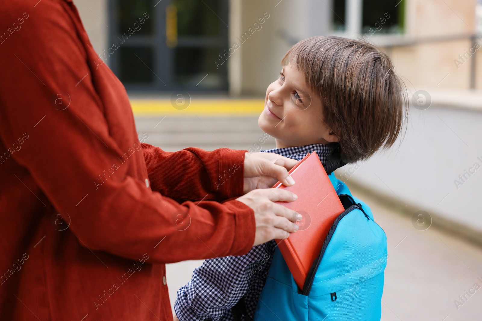 Photo of Mother putting book into son's backpack while taking him to school outdoors, closeup