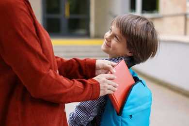 Photo of Mother putting book into son's backpack while taking him to school outdoors, closeup