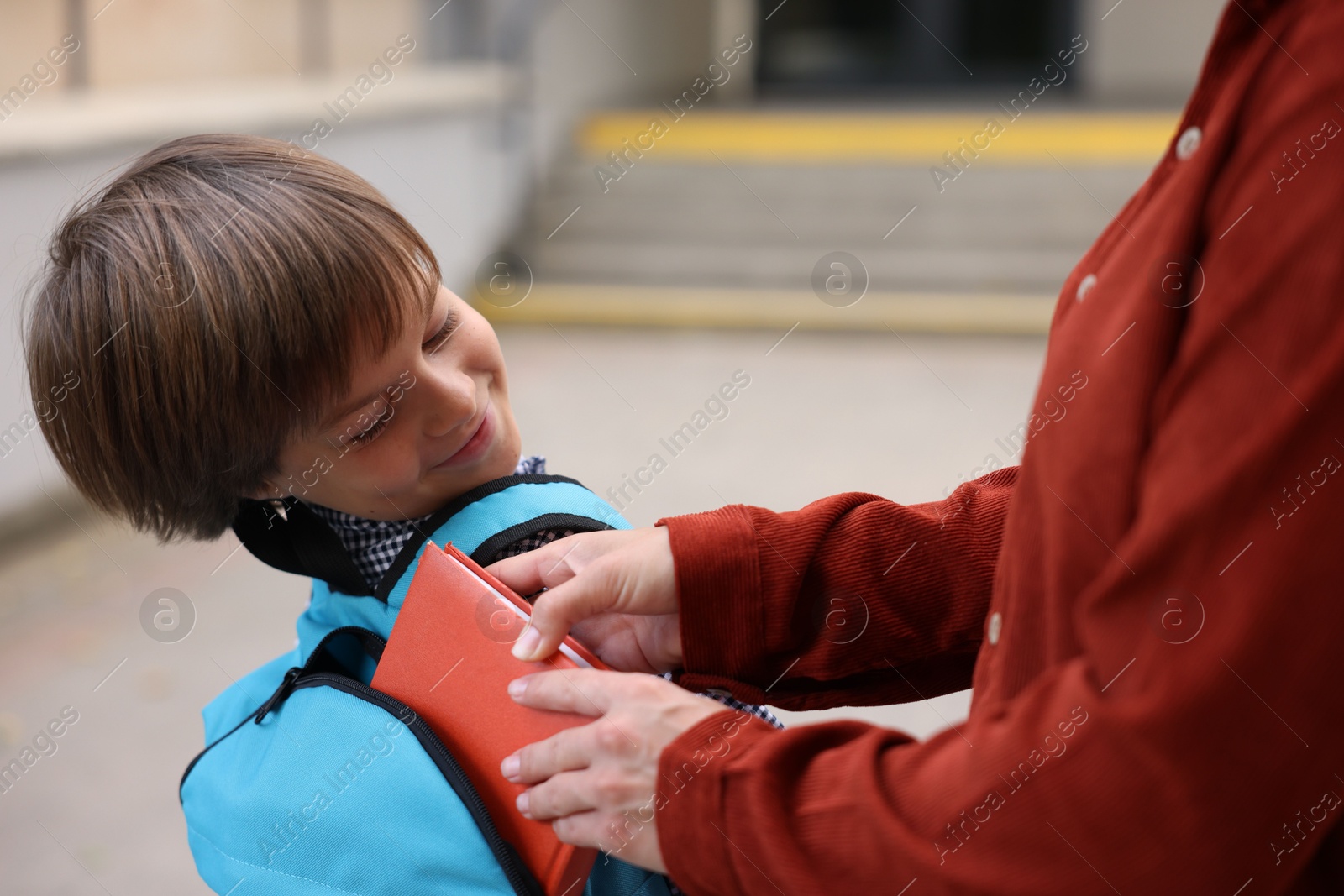 Photo of Mother putting book into son's backpack while taking him to school outdoors, closeup