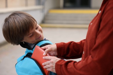 Photo of Mother putting book into son's backpack while taking him to school outdoors, closeup