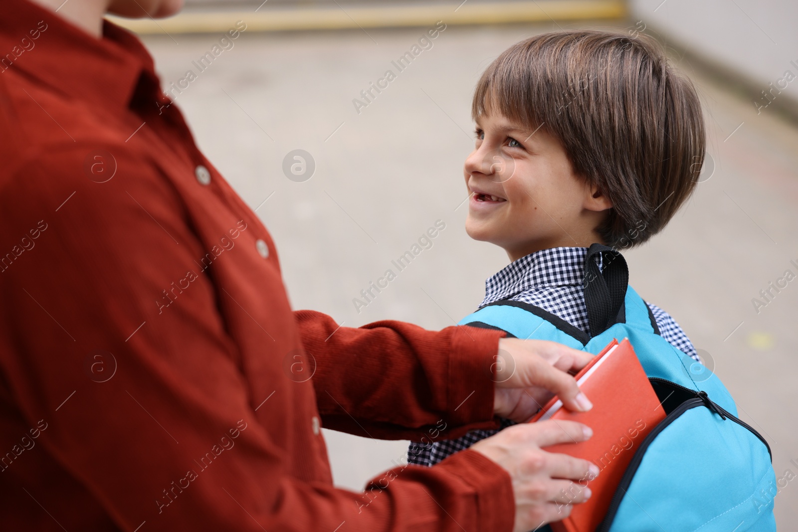 Photo of Mother putting book into son's backpack while taking him to school outdoors, closeup
