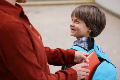 Photo of Mother putting book into son's backpack while taking him to school outdoors, closeup