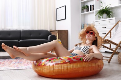 Photo of Happy woman in sunglasses and straw hat with mason jar of juice and inflatable ring having fun on floor at home. Dreaming about vacation at sea