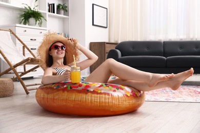 Photo of Happy woman in sunglasses and straw hat with mason jar of juice and inflatable ring having fun on floor at home. Dreaming about vacation at sea