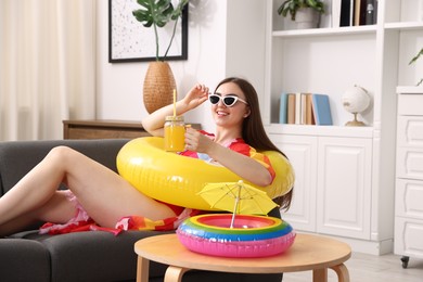 Photo of Happy woman in sunglasses with mason jar of juice and inflatable ring on sofa at home. Dreaming about vacation at sea