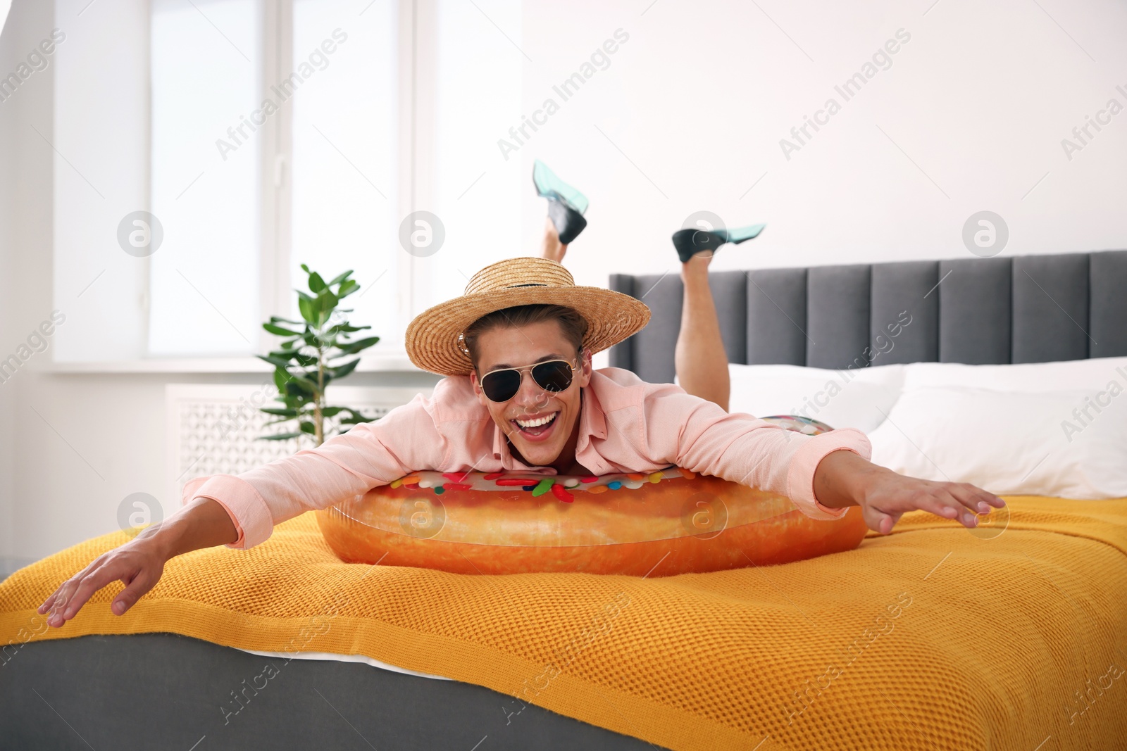 Happy man in straw hat and sunglasses with inflatable ring on bed at home. Dreaming about vacation at sea Photo of Happy man in straw hat and sunglasses with inflatable ring on bed at home. Dreaming about vacation at sea