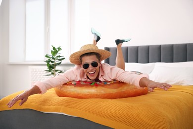 Photo of Happy man in straw hat and sunglasses with inflatable ring on bed at home. Dreaming about vacation at sea