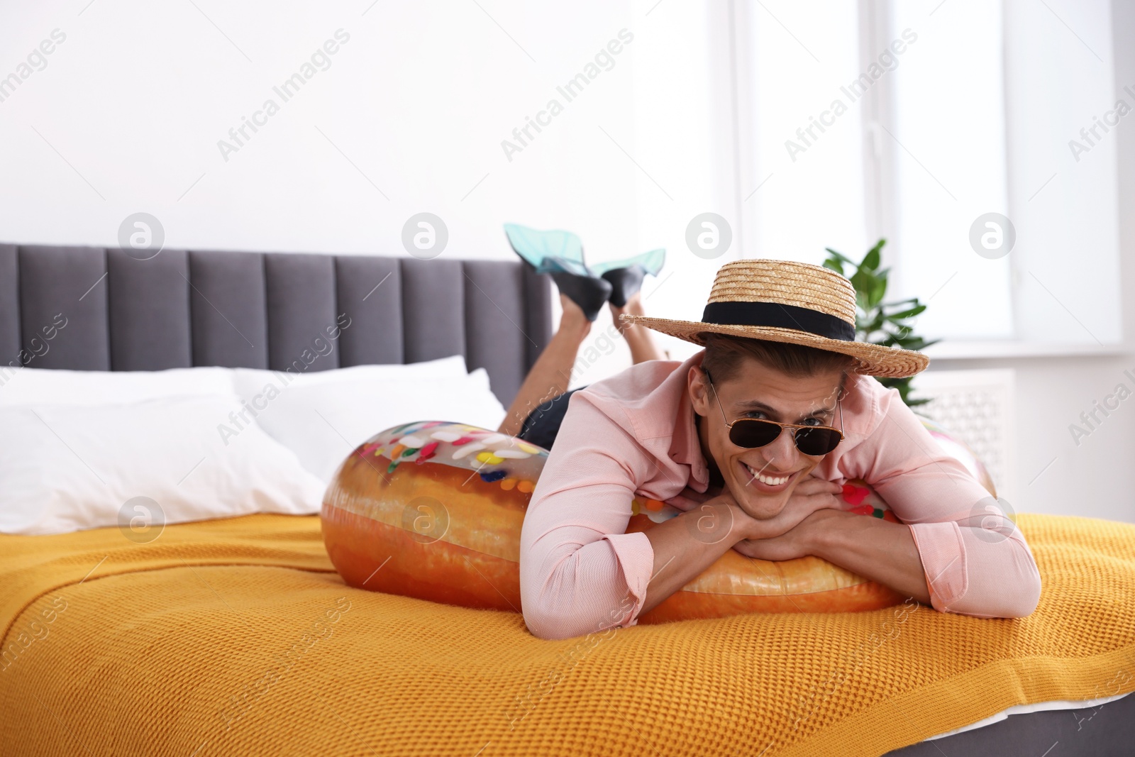 Happy man in straw hat and sunglasses with inflatable ring on bed at home. Dreaming about vacation at sea Photo of Happy man in straw hat and sunglasses with inflatable ring on bed at home. Dreaming about vacation at sea