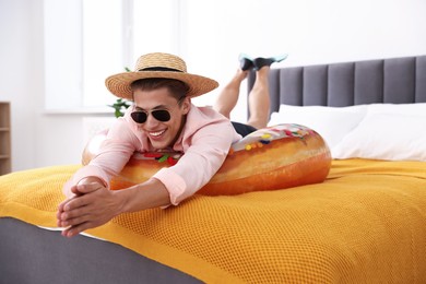 Photo of Happy man in straw hat and sunglasses with inflatable ring on bed at home. Dreaming about vacation at sea